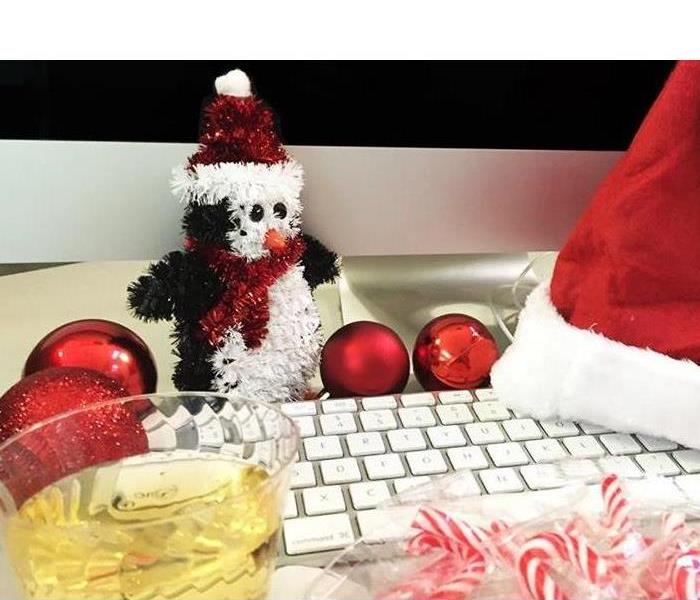 Desktop keyboard with a Santa hat on it, ornaments and candy canes are nearby as well as a yellow beverage in a clear glass