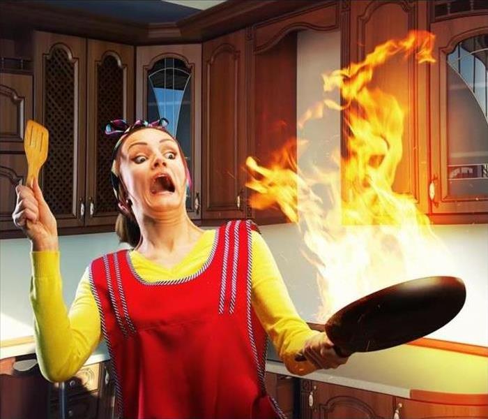A shocked woman in an apron is holding a pan on fire in a kitchen.